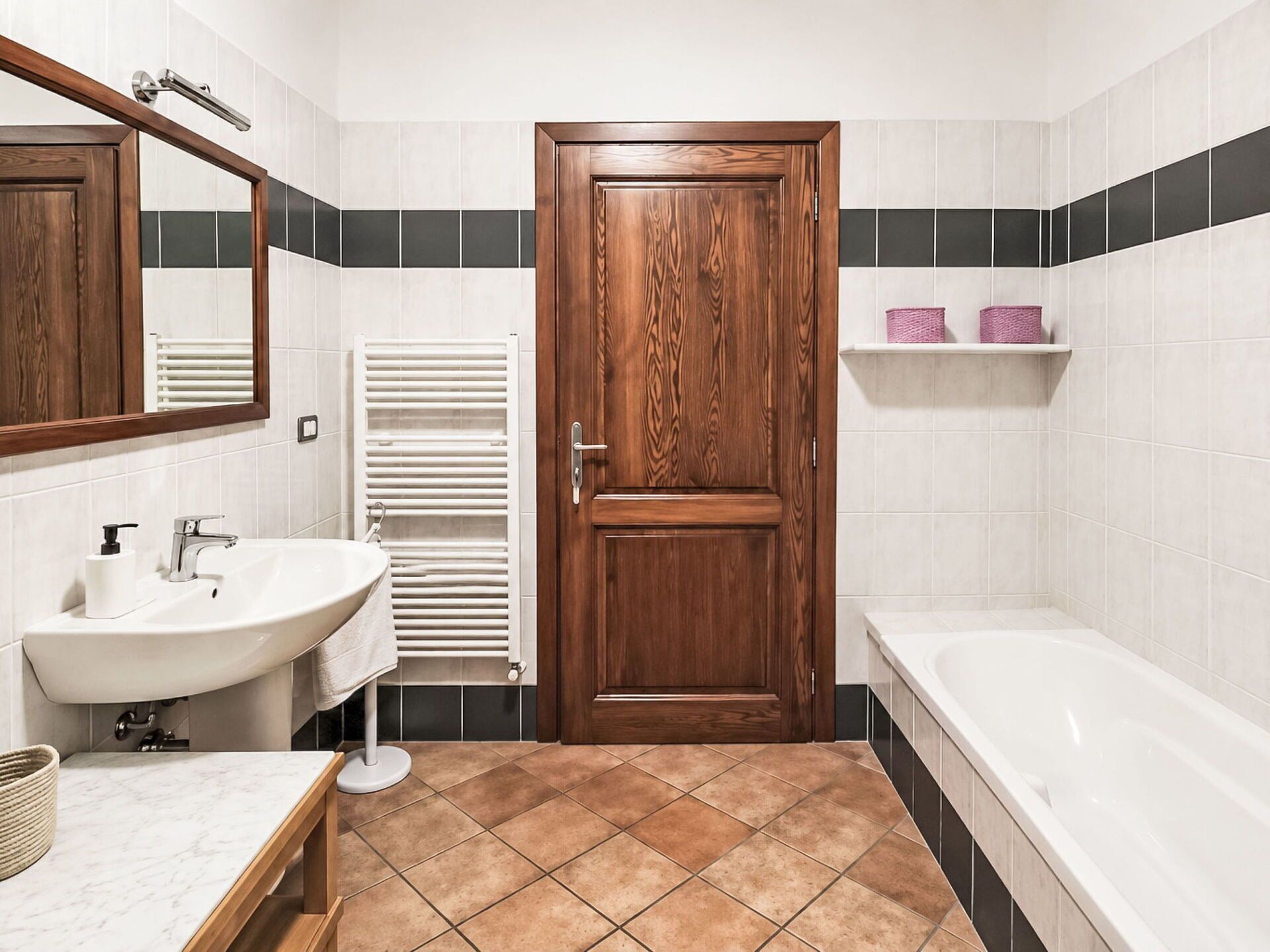 A bathroom with a wooden door, white tiles, a white sink, a white bathtub, and a towel rack.