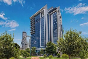 Un edificio moderno de gran altura con fachada de vidrio, rodeado por un parque con árboles y una noria.