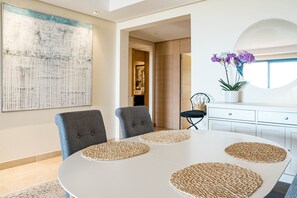 A dining area with a round table, grey upholstered chairs, woven placemats, a large abstract painting, a potted orchid, and a white cabinet with drawers. - BVQ - Stunning Sea & Golf View Terrace (Benahavis)