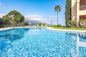 A swimming pool with a tiled edge, surrounded by a fence, palm trees, and a building with a balcony. - BVQ - Stunning Sea & Golf View Terrace (Benahavis)
