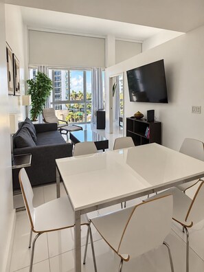 Een moderne woonkamer met een bank, een eettafel met stoelen, een flatscreen televisie aan de muur en een groot raam met uitzicht op palmbomen en gebouwen.