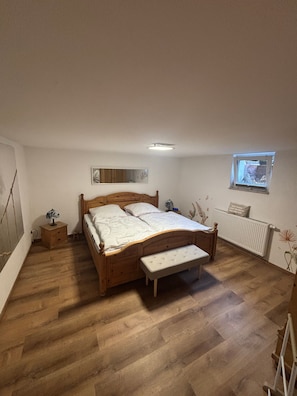 A bedroom with a wooden bed, a nightstand, a bench, a radiator, a window, and a wall-mounted mirror. - Cozy 45 m² Apartment in the Vineyards (Bad Kreuznach)