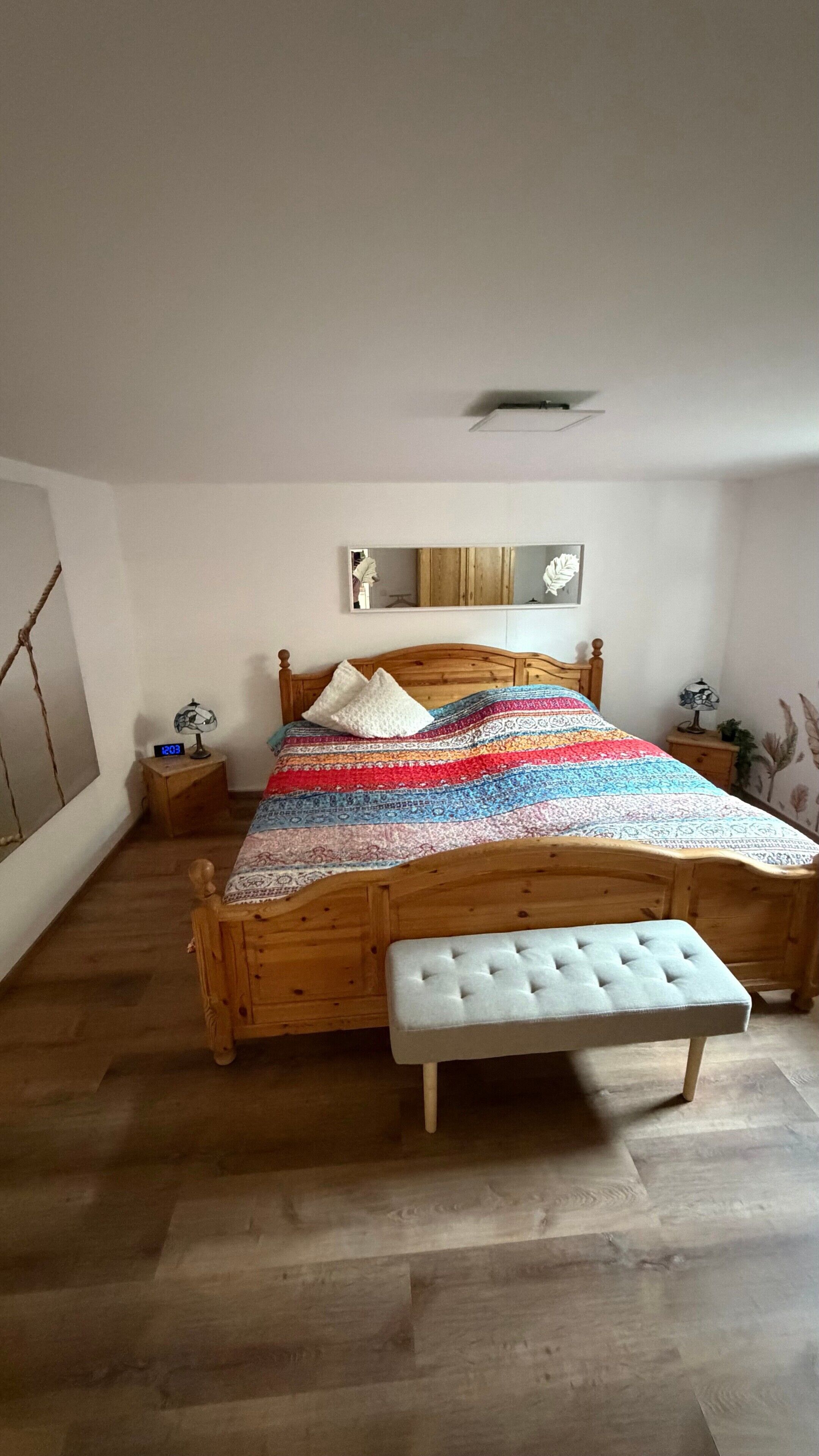 Ein Schlafzimmer mit einem Holzbett, einer Bank, einem Nachttisch mit Lampe und einem Wandspiegel.