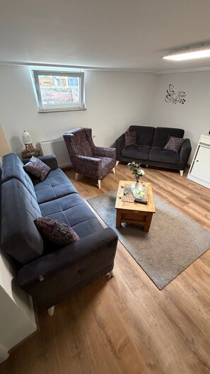 Ein Wohnzimmer mit einem blauen Sofa, einem gemusterten Sessel, einem dunklen Sofa und einem Couchtisch aus Holz mit einer Vase und Fernbedienungen.