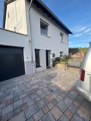 A two-story building with a garage, a small front porch, and a paved driveway. - Cozy 45 m² Apartment in the Vineyards (Bad Kreuznach)