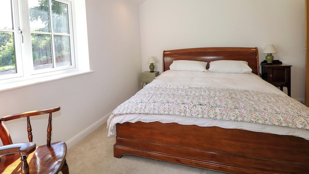 A bedroom with a wooden bed, a window, a chair, and a nightstand with a lamp. - Keepers Cottage (Haslemere)