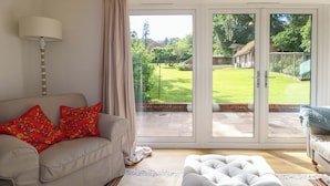 A living room with a sofa, ottoman, and a view through a sliding glass door to a garden. - Keepers Cottage (Haslemere)