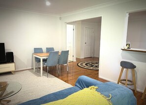 Una sala de estar con un sofá, una mesa de comedor con sillas, un televisor y un pequeño taburete.