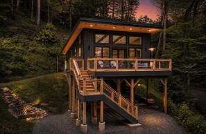 A modern cabin with a wooden deck, a staircase, and a large window overlooking a forested area. - The Acosta (Maggie Valley)