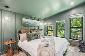 A bedroom with a bed, pillows, a bedside table with a clock, a hanging light, and windows with a view of greenery. - The Acosta (Maggie Valley)