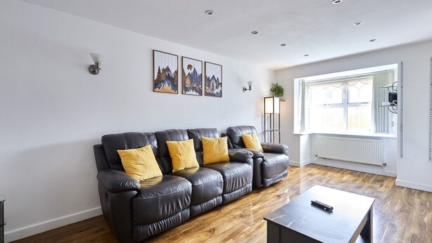 A living room with a black leather sofa, wooden flooring, a small table, and a window with blinds. - Host & Stay | Urban Nest LIV (Liverpool)