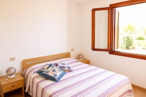 A bedroom with a bed, a nightstand, a bedside lamp, a window with a wooden frame, and a view of greenery outside. - Residence Domenique | Apartment on 2 levels (Tanaunella)