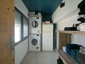 A compact laundry room with two stacked washing machines, a small refrigerator, and a shelf with folded towels.