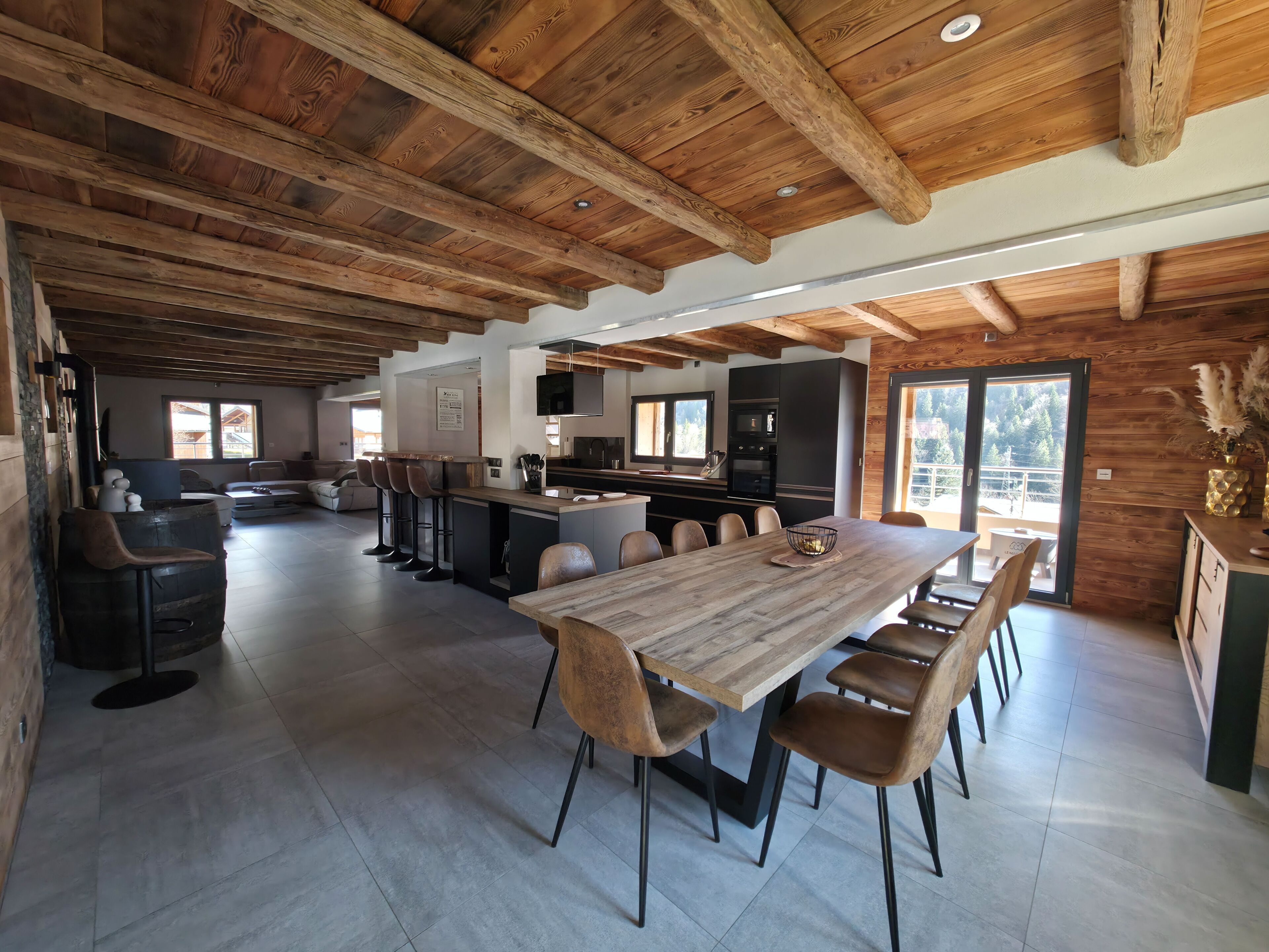 Un vaste espace de vie doté d’un plafond en bois, d’une table à manger avec des chaises, d’un coin cuisine et offrant une vue sur l’extérieur.