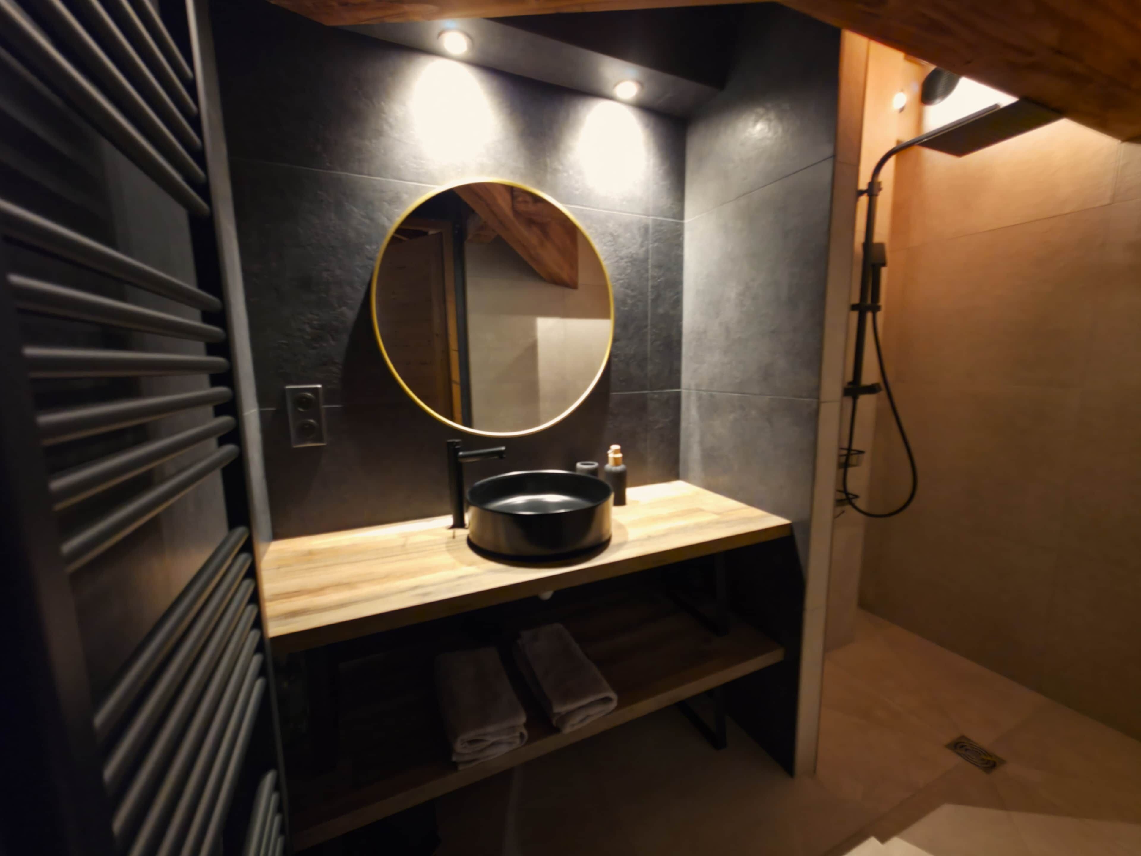 Une salle de bain avec un meuble-lavabo en bois, un miroir rond, un lavabo noir et un espace douche.