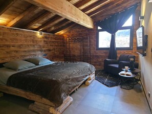 A cozy bedroom with a wooden headboard, a bed with pillows, a chair, a small table with a cup and book, and a window with curtains.