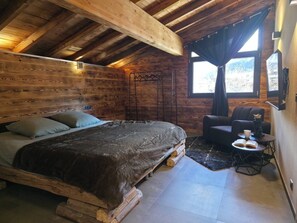 A rustic wooden room with a bed, a chair, a small table with a cup and book, and a window with curtains. - Vrbo Property (Bozel)