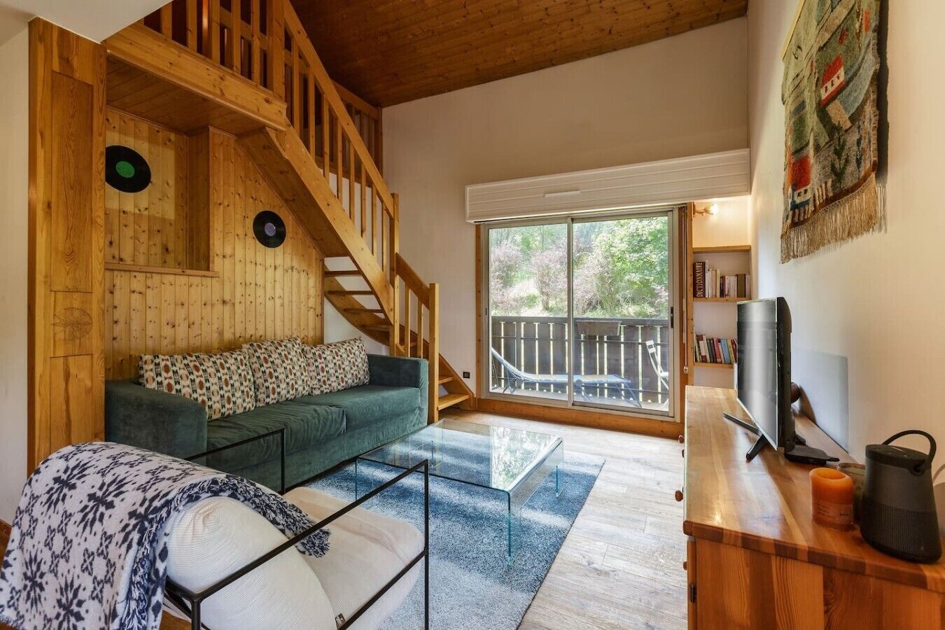 A wooden interior with a staircase, a green sofa, a glass coffee table, and a TV on a wooden stand.