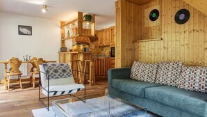 Ein gemütliches Wohnzimmer mit einer Couch und einem Sessel, Holzwänden und einer Küchenzeile mit Holzausstattung und einer hängenden Pflanze.