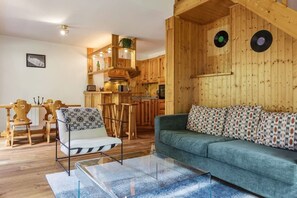 Ein gemütliches Wohnzimmer mit einer Couch und einem Sessel, Holzwänden und einer Küchenzeile mit Holzausstattung und einer hängenden Pflanze.