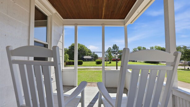 Une véranda avec des chaises en bois peintes en blanc, un plafond en bois et une vue sur une pelouse verdoyante et des bâtiments situés au loin.