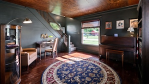 Une pièce avec un plafond en bois, un piano, un bureau avec une lampe, une chaise, une grande moquette et un escalier.