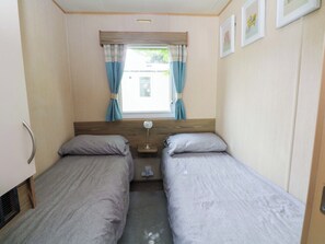A compact bedroom with two single beds, a window with curtains, a bedside table, and a wall-mounted heater. - Kensington 49 (Blackpool)