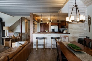 Een ruime woonkamer met een houten plafond, een accentmuur van steen, een eettafel met stoelen en een bar met krukken.