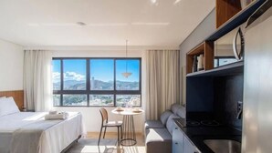 A hotel room with a bed, a dining table, a sofa, a kitchenette, and a large window with a city view.