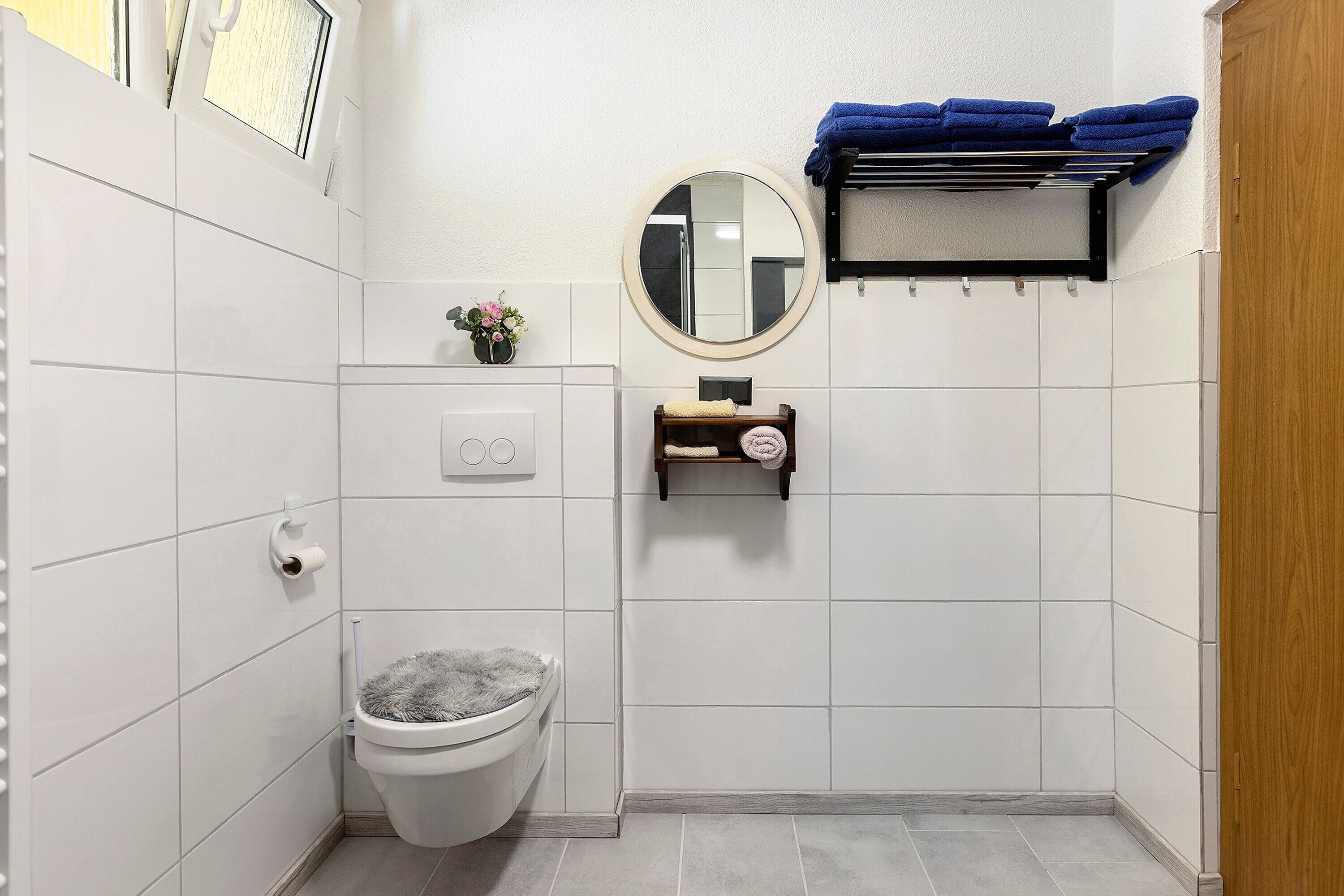 Un baño con azulejos blancos, un inodoro, un estante pequeño con un espejo y un perchero con toallas azules.