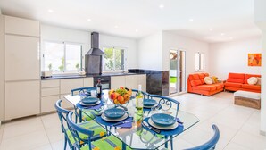 A modern kitchen with a dining area, featuring a glass table, blue chairs, and a fruit bowl. - Elegant Villa w/ Heatable Pool and BBQ (Albufeira)
