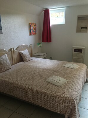 Une chambre à coucher comprenant un lit, une table de chevet, une lampe, une fenêtre avec un rideau et un petit meuble.