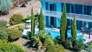 A two-story house with a swimming pool, surrounded by greenery and tall cypress trees.