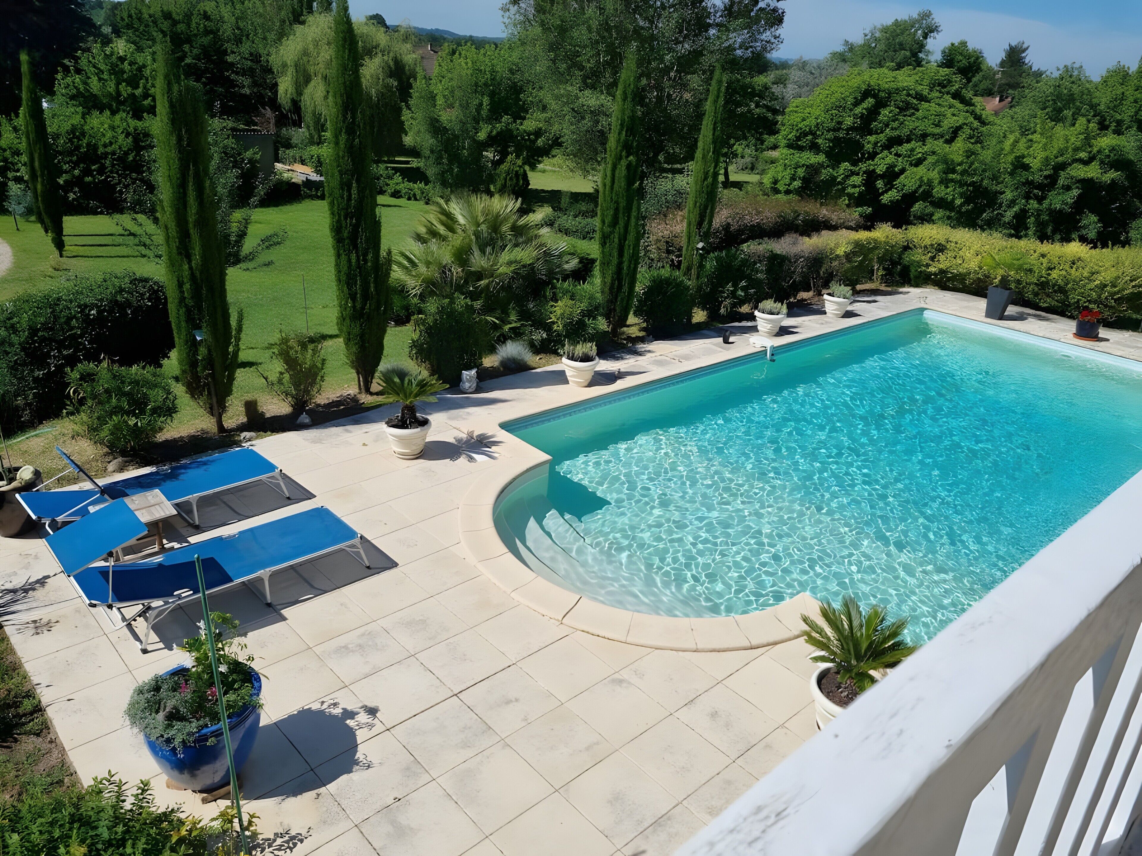 A rectangular swimming pool with a curved edge, surrounded by a tiled deck with lounge chairs and potted plants.. Ground-Floor Apartment for 2-3 Guests in Périgord Pourpre with Pool