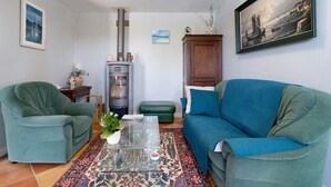 A living room with two green armchairs, a blue sofa, a glass coffee table, a patterned rug, a fireplace, and a painting on the wall.