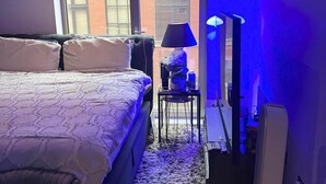 Ein Schlafzimmer mit einem Bett, einem Nachttisch mit Lampe und Blick auf ein Backsteingebäude durch ein Fenster.