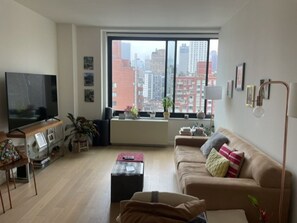 A living room with a large window, a flat-screen TV, a sofa, a coffee table, and a floor lamp. - Beautiful New York City apartment, prime location and access to a roof deck (New York)