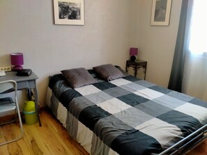 En seng med ternet sengetæppe, et skrivebord med lampe, en stol og et lille bord med lampe.