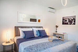 Un dormitorio con cama, mesitas de noche, escritorio, espejo y aire acondicionado de pared.