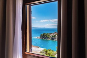 A coastal view through a window with curtains, a clear sky, and a distant shoreline with buildings. - Villa Kadena (Selca, Brač)