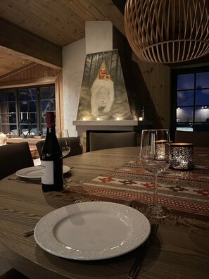 Una mesa de comedor con una botella de vino, copas y platos, una chimenea con un cuadro de un oso, y una alfombra estampada.