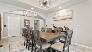 Uma sala de jantar com uma grande mesa de madeira, seis cadeiras, um lustre e um quadro na parede.