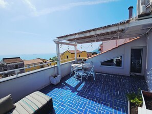 Une terrasse sur le toit, avec un sol carrelé de bleu, une table avec des chaises, une petite plante en pot et une vue sur la mer et les bâtiments.