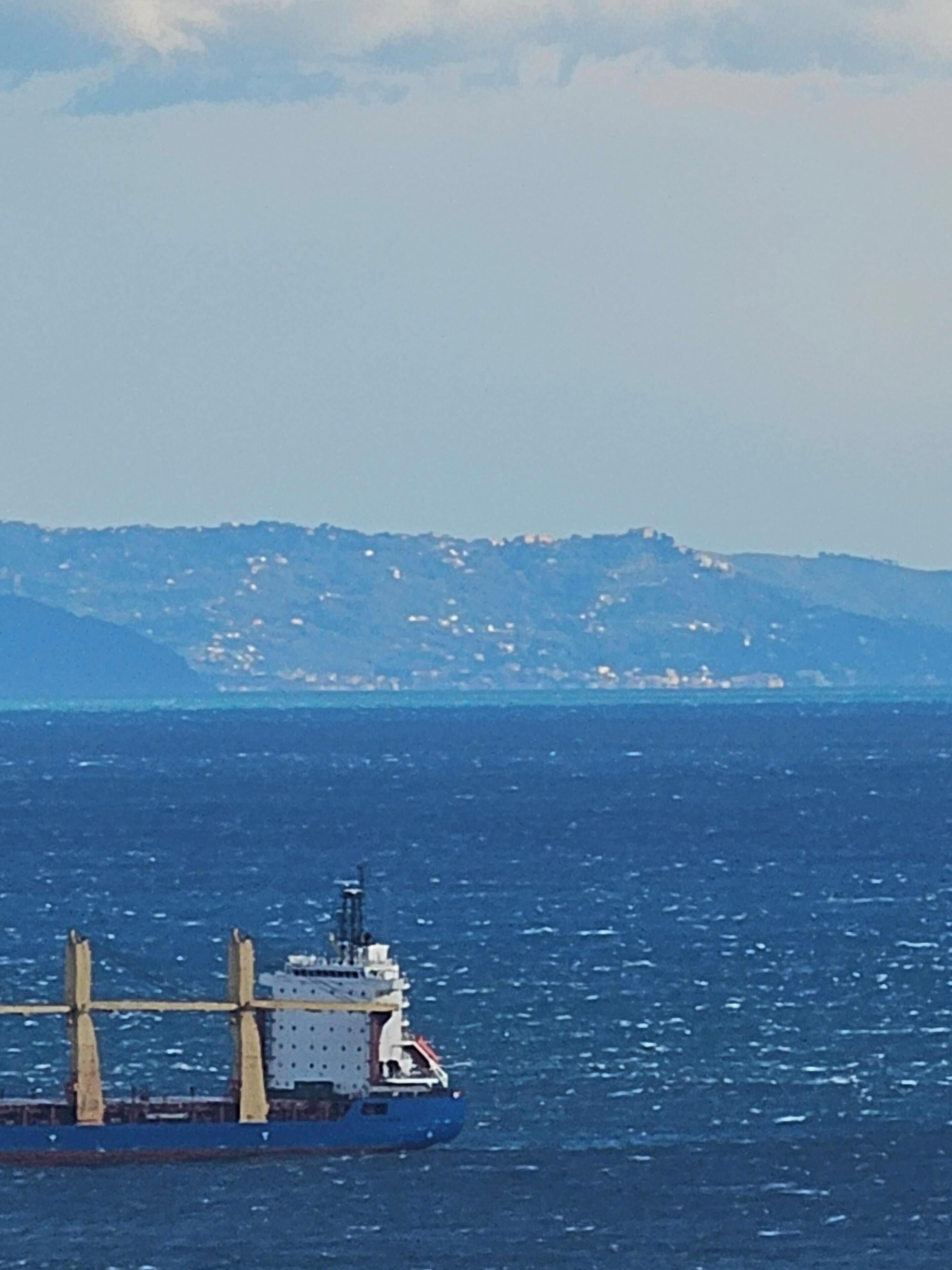 Ein Frachtschiff auf dem Ozean vor einer Küstenlandschaft mit Bergen im Hintergrund.