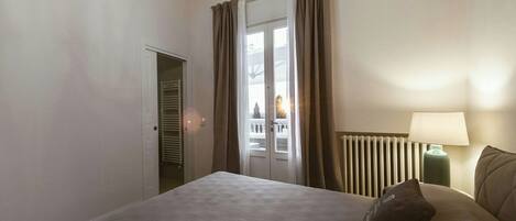 Un dormitorio con cama, mesita de noche con lámpara, ventana con cortinas y radiador.