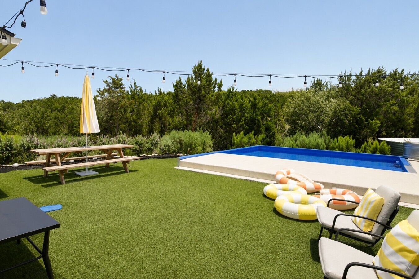 A backyard with a swimming pool, artificial grass, a picnic table, lounge chairs, and string lights.