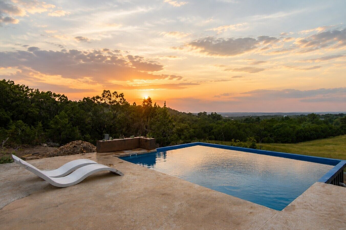 A swimming pool with a curved edge, surrounded by a concrete deck, set against a backdrop of trees and a sunset.