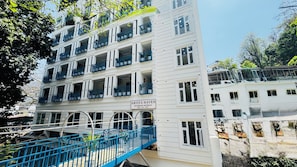 A multi-story hotel with balconies, a sign indicating it is the Arista Haven Riverside Retreat, and a blue bridge leading to the entrance. - Arista Haven Hotel (Narendranagar)