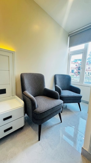 A modern room with two grey armchairs, a white bedside table, and a large window with a view of buildings. - Arista Haven Hotel (Narendranagar)