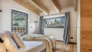 A bedroom with a bed, a window with a view of a house and trees, wooden ceiling beams, and a door. - Rock Art Luxury Attic (Ponte di legno)